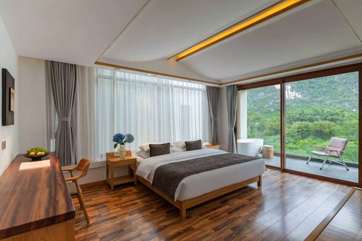 Ein modernes Schlafzimmer mit einem großen Bett, Holzboden, einem Schreibtisch und Blick auf einen üppigen Wald durch große Fenster.