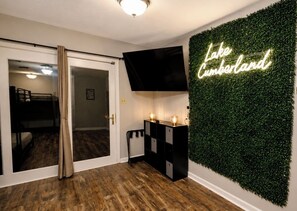 A room with a wooden floor, a sliding door, a black console table with two lamps, and a neon sign on the wall. - Private Indoor Pool + Slide Sleeps 18+ Lake Cumberland (Somerset)