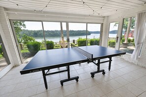 Una mesa de ping pong en una habitación con ventanas amplias que dan a un lago y vegetación.
