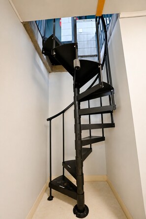 Una escalera espiral de metal negro en una esquina de una habitación, con una pared blanca y una puerta azul al fondo.