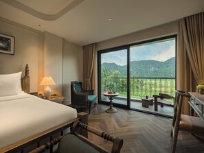 Habitación de hotel con una cama grande, una silla, una mesita con fruta y un balcón con vistas a zonas verdes y montañas.