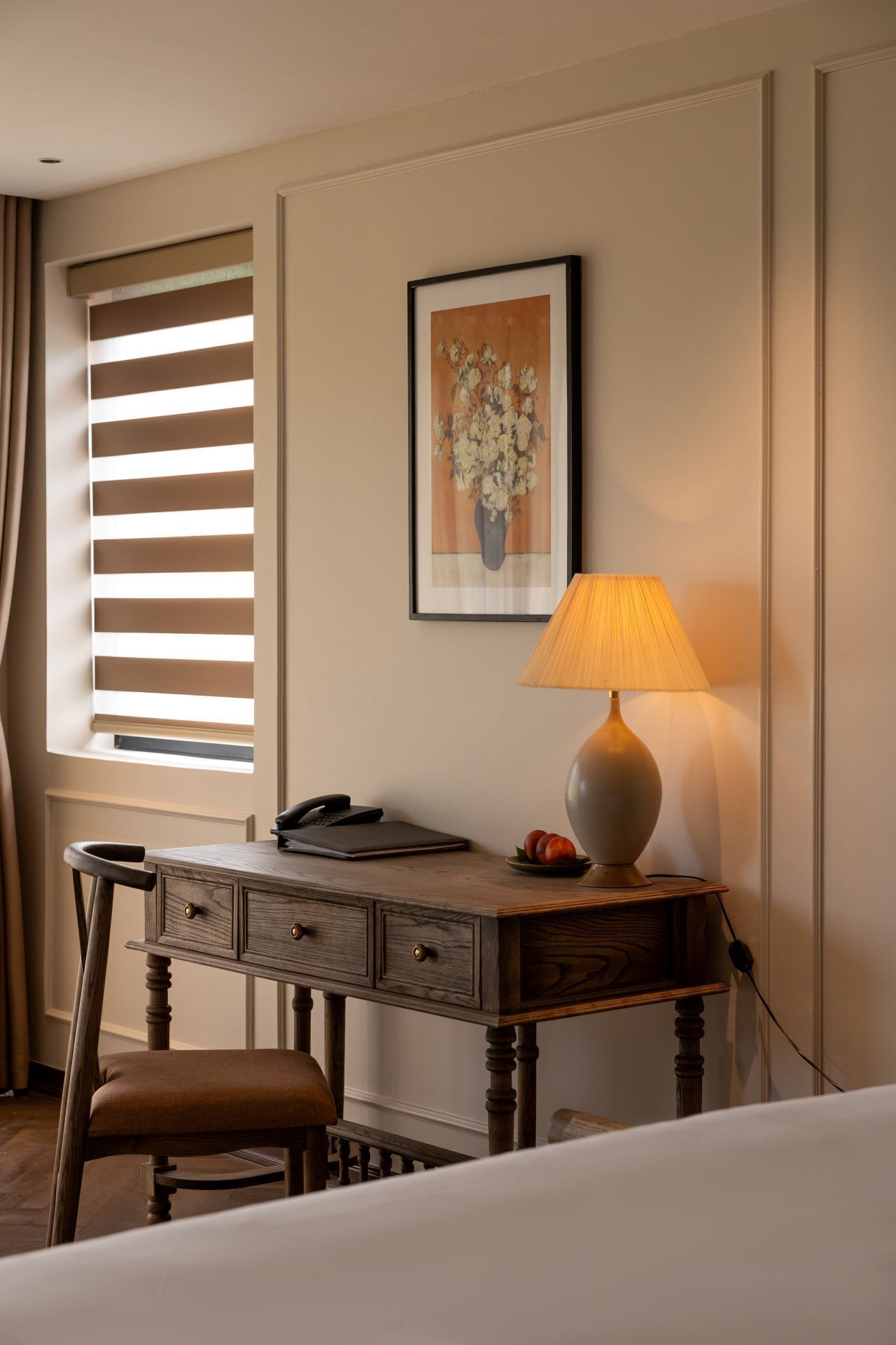 A hotel room with a wooden desk, a lamp, a framed picture, and a window with blinds.