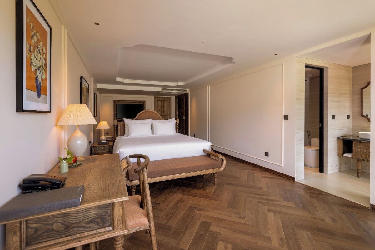 Habitación de hotel con cama, muebles de madera, escritorio y baño visible a través de una puerta abierta.