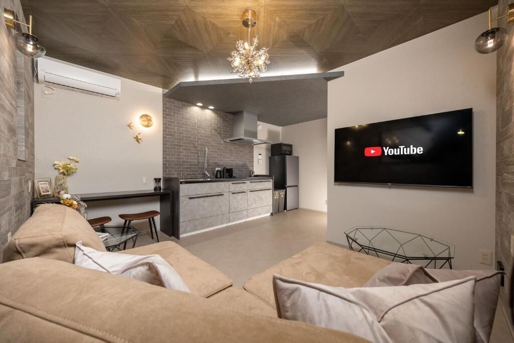 Uma sala de estar moderna com um sofá, uma televisão na parede e uma cozinha ao fundo.
