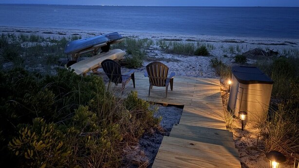 A wooden deck with chairs and a kayak, leading to a beach with a view of the ocean.