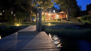 A wooden dock leading to a house with a lit porch, surrounded by trees and a body of water.