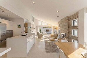 A modern kitchen with a central island, a dining area with a wooden table, and a living space with a fireplace. - The Lumiere House -Elegance in Mt Maunganui (Mount Maunganui)
