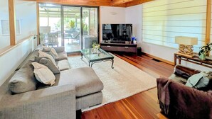 Ein modernes Wohnzimmer mit Holzboden, einem Ecksofa, einem Flachbildfernseher und einer Glastür, die zu einem Außenbereich führt.