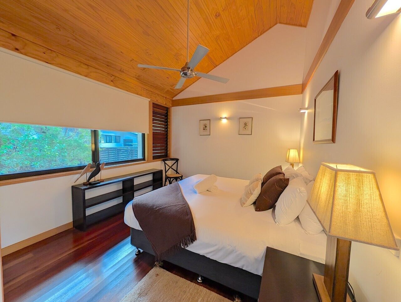 A bedroom with a bed, bedside lamps, a ceiling fan, a window with blinds, and a desk with a chair.