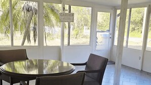 Un espacio al aire libre cubierto con una mesa redonda y sillas, un letrero que dice 'GARDEN' y grandes ventanales con vistas a palmeras.