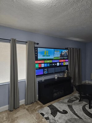 A living room with a flat-screen TV mounted on the wall, a black entertainment unit below it, and a patterned rug on the floor. - Walk to Casino, Fairground, Drive to beaches, King Bed (Tampa)