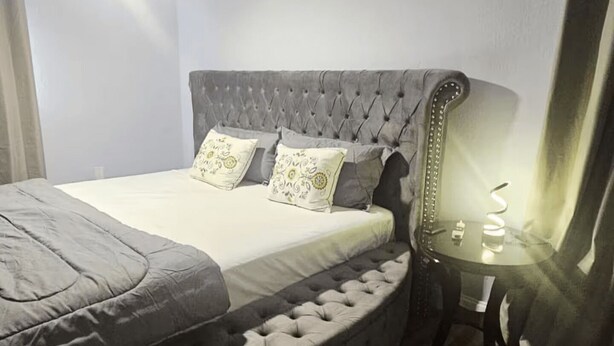 Un dormitorio con cabecera capitonada, cama con ropa de cama gris, mesita de noche con lámpara y ventilador de techo.