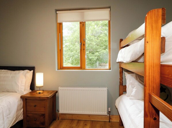 Una habitación con una cama, una mesita de noche con lámpara, un radiador y una ventana con vista a árboles.
