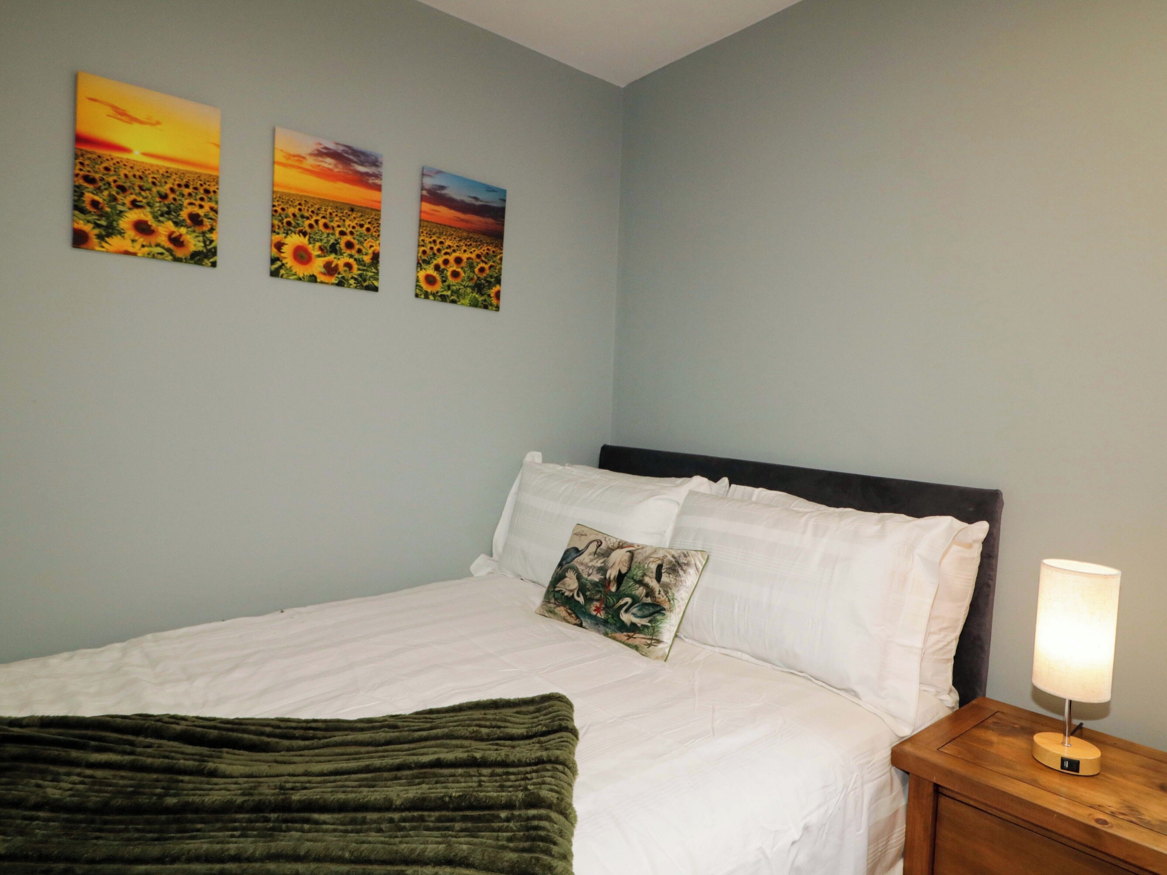 A bedroom with a bed, a nightstand, a lamp, and three wall-mounted paintings of sunflowers.