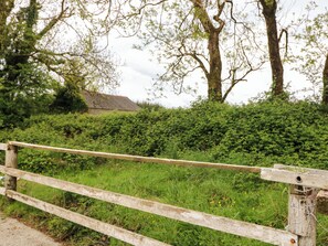 A rustic wooden fence, a hedge, and a building with a thatched roof in the background.