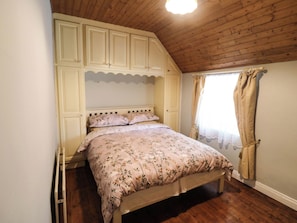 Un dormitorio con techo de madera, cama con ropa de cama floral, armario y ventana con cortinas.