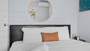 Una cama con ropa blanca y un almohadón marrón, un espejo redondo en la pared y un cuadro abstracto azul.
