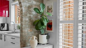 Una cocina con pared de ladrillo, gabinetes blancos, un lavamanos, una cafetera, una planta en maceta, un florero y una ventana con persianas.