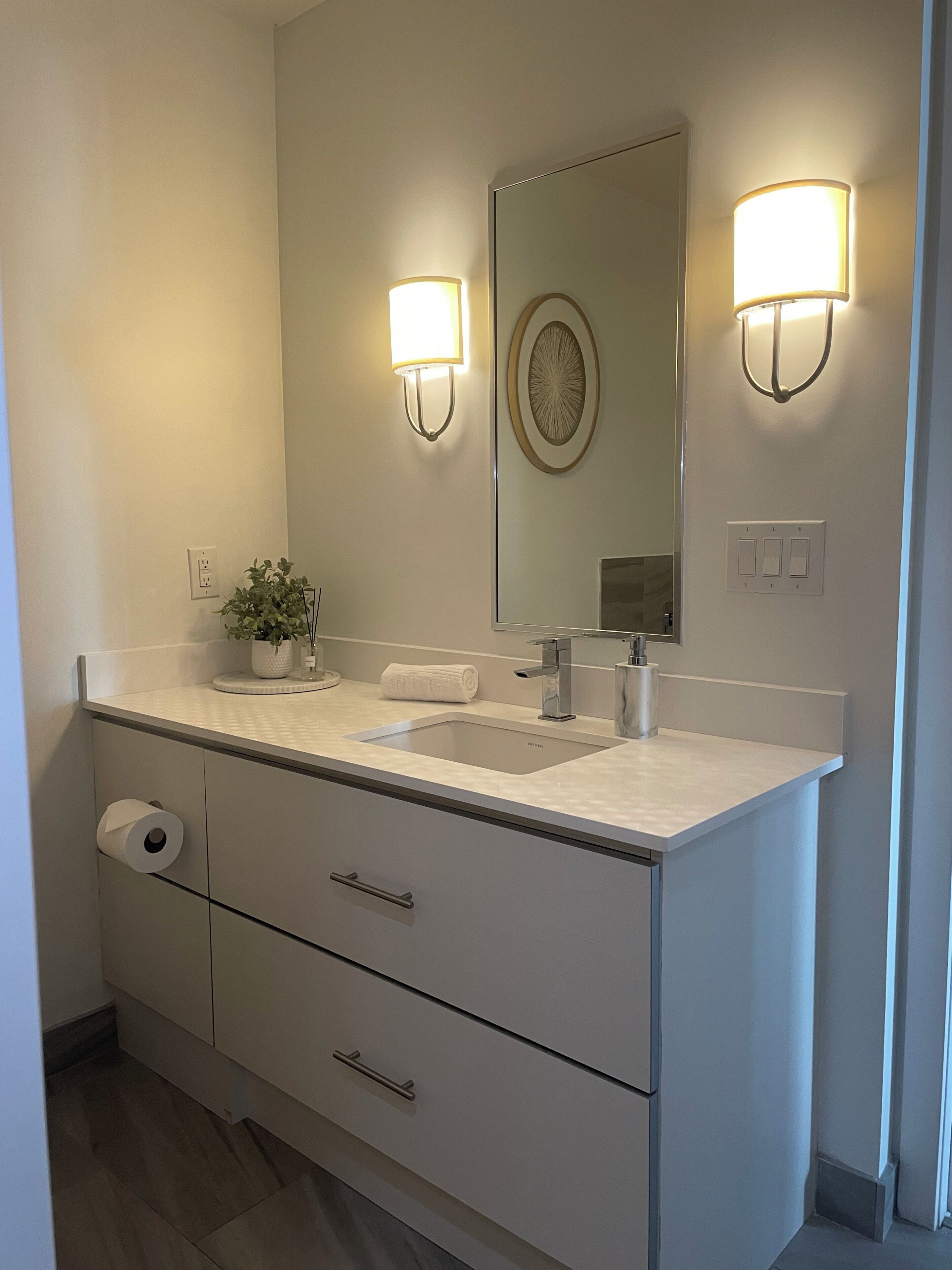 A modern bathroom with a sink, mirror, wall-mounted lights, a towel, and a toilet paper holder.