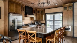 Une cuisine avec des meubles en bois, un réfrigérateur en acier inoxydable, un grand îlot central avec des tabourets, et une fenêtre avec des stores.