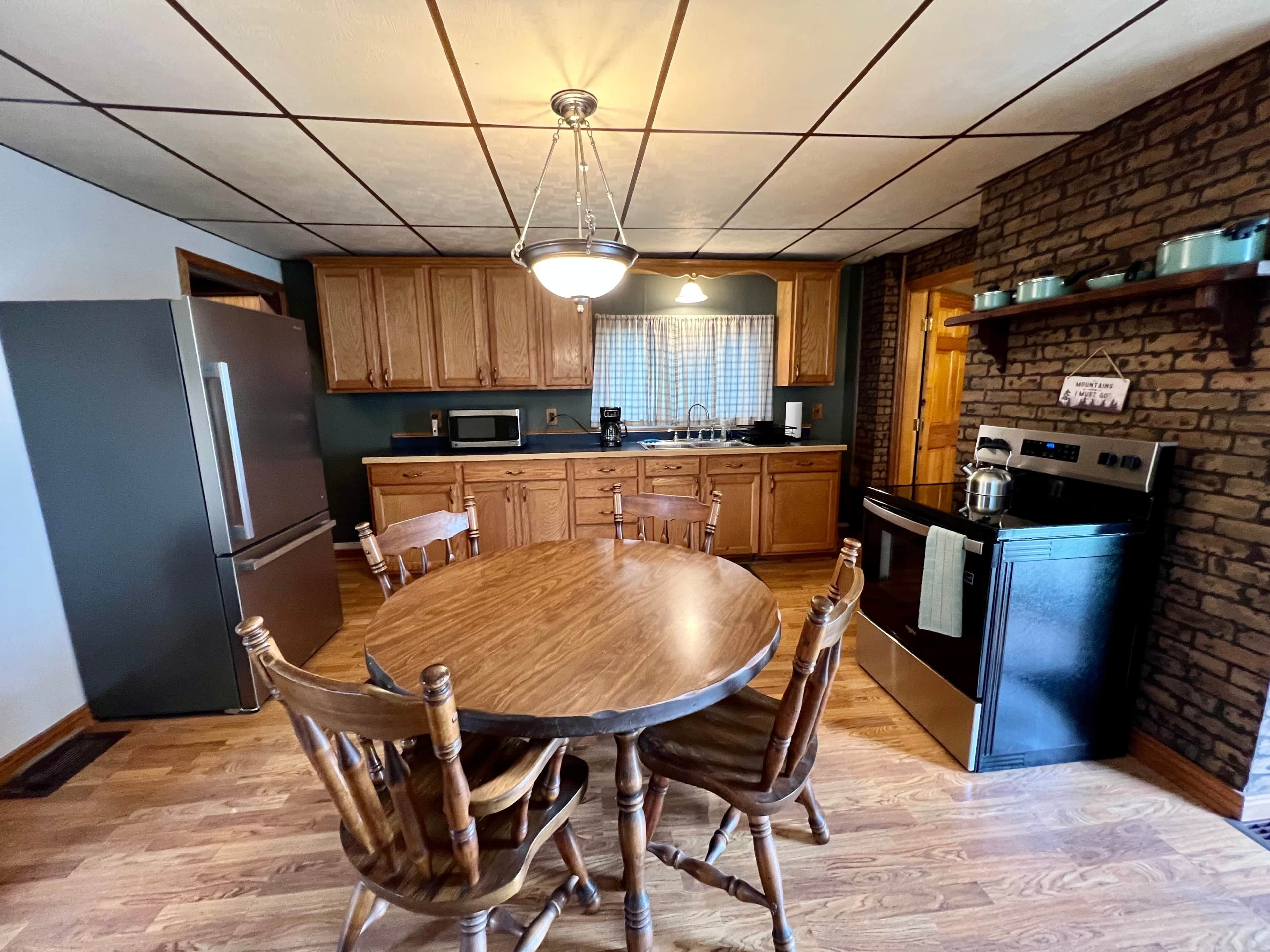 A kitchen with wooden cabinets, a dining table with chairs, a refrigerator, a microwave, a stove, and a hanging sign.