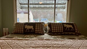 Un lit avec une couverture à carreaux et deux oreillers ornés de motifs d’élans, une fenêtre donnant sur des maisons et une lampe sur la table de chevet.