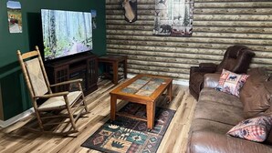 A rustic living room with a wooden floor, a leather sofa, a wooden rocking chair, a glass-top coffee table, a television, and a ceiling fan. - The Buck Lodge | Arcade • Firepit (Wheeling)
