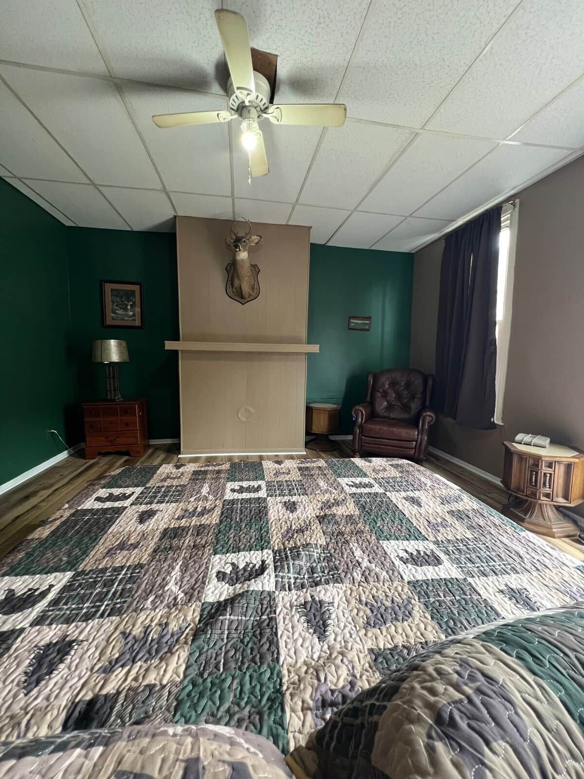 A room with a ceiling fan, a mounted deer head, a quilted bedspread, a leather chair, and a small wooden cabinet.