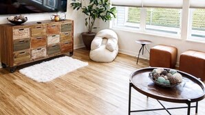 Un salon moderne avec un plancher en bois, un grand téléviseur à écran plat, un meuble en bois avec des tiroirs, une plante en pot, un pouf blanc et une table basse ronde.