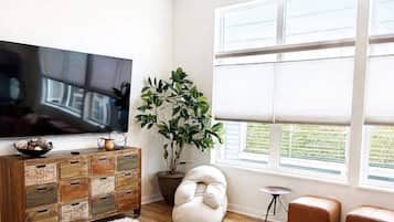 A modern living room with a wooden floor, a large flat-screen TV, a wooden cabinet with drawers, a potted plant, a white pouf, and a round coffee table.