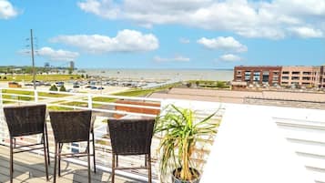 A rooftop terrace with wicker chairs, a potted plant, and a view of a beach and parking lot.