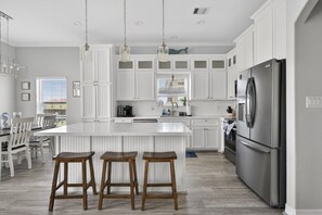 Une cuisine moderne avec des meubles blancs, un grand îlot central doté de tabourets, un réfrigérateur en acier inoxydable et des luminaires suspendus.