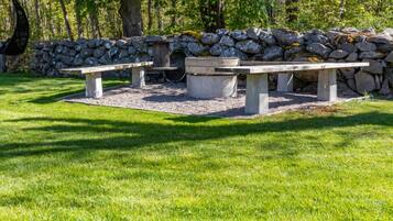 Un muret de pierres, un foyer et deux tables de pique-nique dans un espace herbeux.