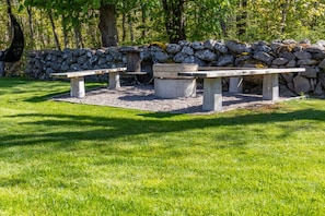 A stone wall, a fire pit, and two picnic tables in a grassy area.