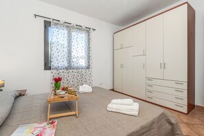 A bedroom with a bed, a wardrobe, a window with curtains, a bedside table with a tray of items, and a painting on the floor. - Le Dimore Del Sole, Olbia, Italy (Olbia)