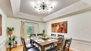 Una sala da pranzo con un tavolo in legno apparecchiato per sei persone, circondato da sedie, un lampadario moderno, piante in vaso e un grande quadro astratto appeso alla parete.