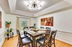 A dining room with a wooden table set for six, surrounded by chairs, with a modern chandelier, potted plants, and a large abstract painting on the wall. - Spacious Home-Game Room - Private Pond - Sleeps 16 (Midland)