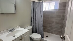 Un bagno con lavandino, water e zona doccia, tutti in bianco, uno specchio illuminato e una finestra con persiane.