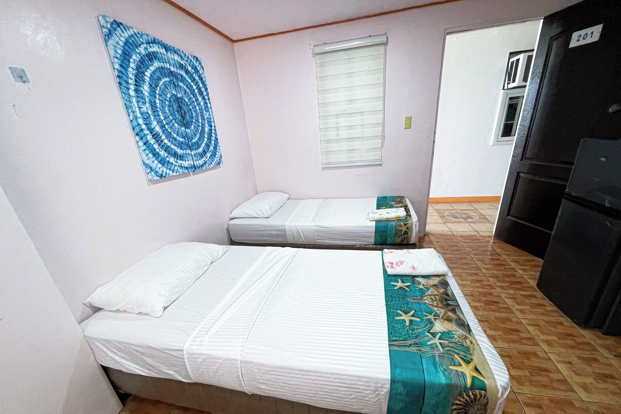 A room with two beds, a wall art piece, a window with blinds, and a kitchen area.