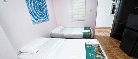 A room with two beds, a wall art piece, a window with blinds, and a kitchen area.