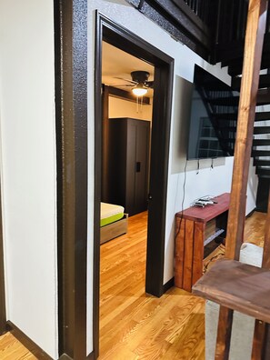 A narrow hallway with a wooden floor leading to a room with a bed and a ceiling fan. A staircase is visible to the right.