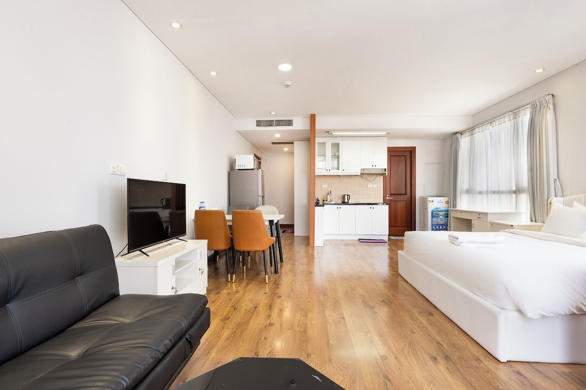 A modern hotel room with a black leather sofa, a flat-screen TV, a dining area with orange chairs, a kitchenette, and a large bed.