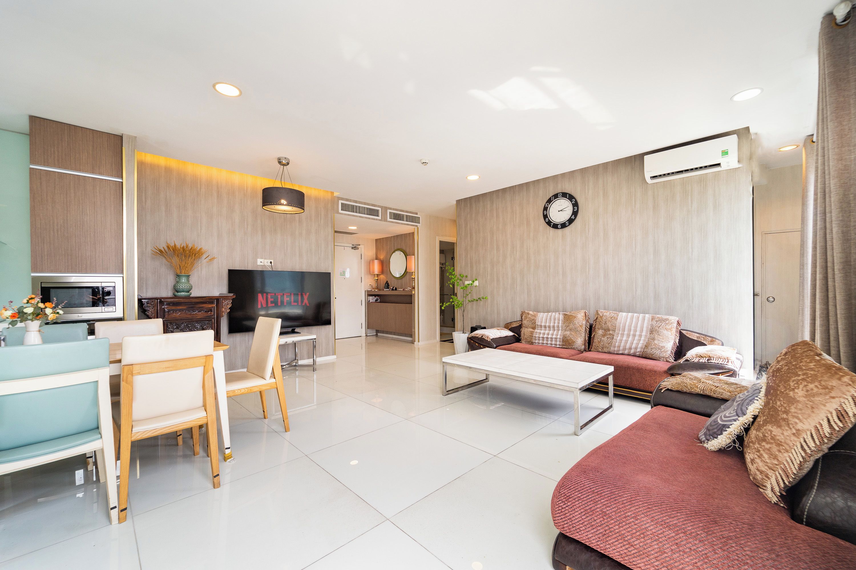 A modern living room with a sofa, coffee table, and TV. There is a dining area with a table and chairs. The room has a wooden floor and a clock on the wall.