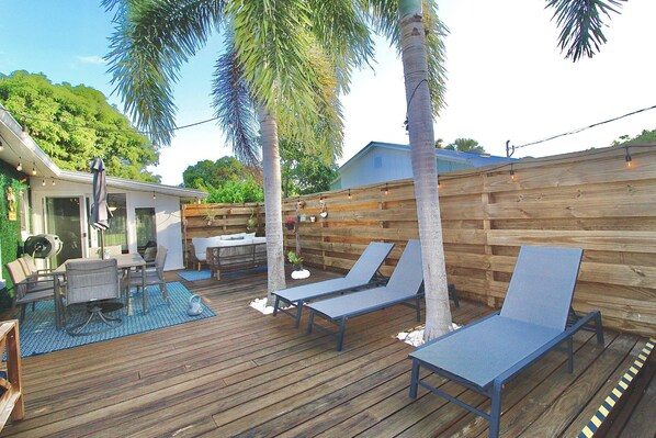 Um deck de madeira com espreguiçadeiras, uma piscina e um alpendre com mesa e cadeiras.