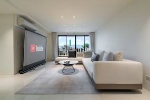 Ein modernes Wohnzimmer mit großem Flachbildfernseher, einer Couch, einem Couchtisch und Blick auf eine Skyline durch große Fenster.