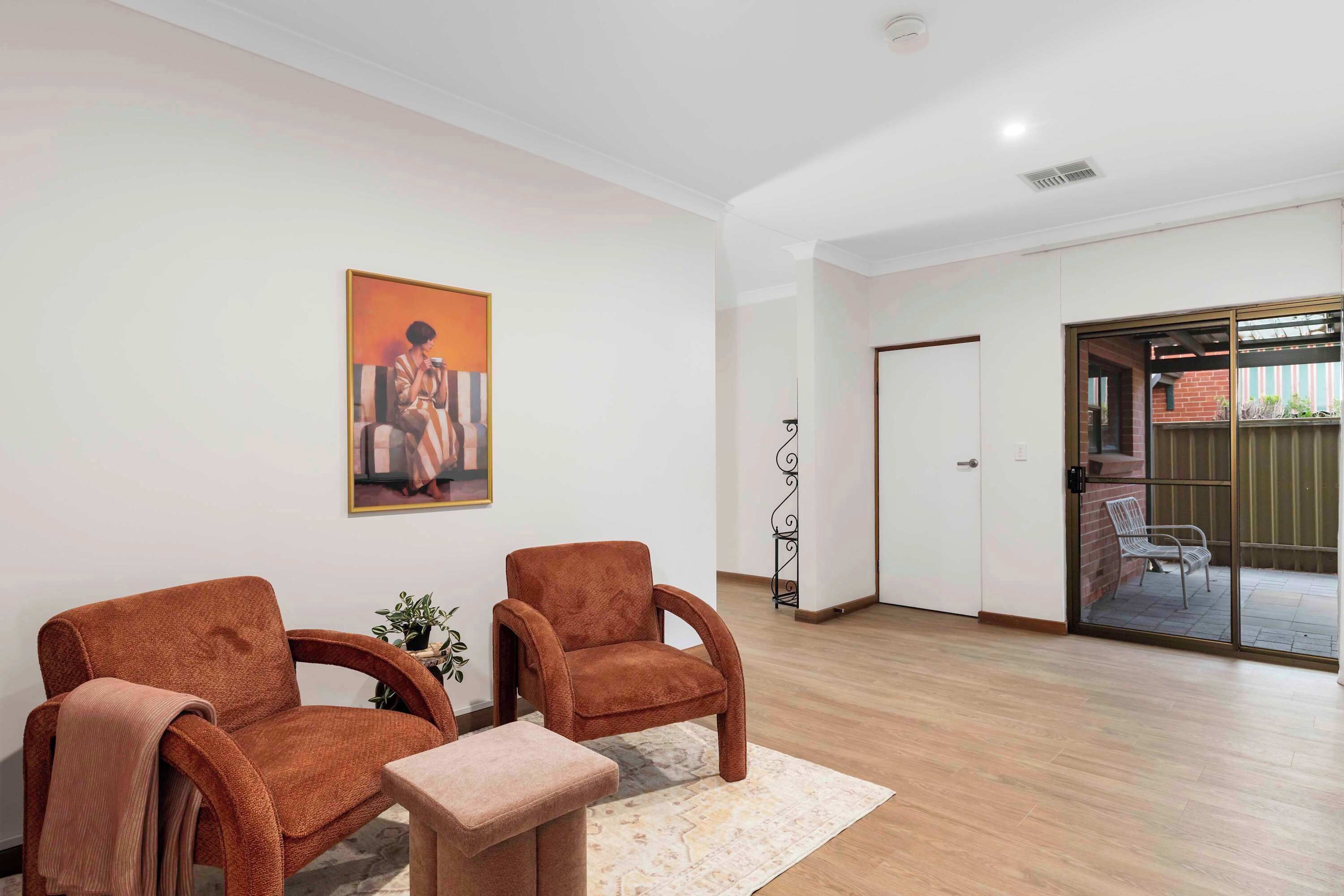 A room with two brown armchairs, a small table, a painting on the wall, and a sliding glass door leading to an outdoor area.