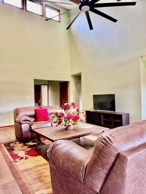 A living room with a brown leather sofa, a wooden coffee table with a vase of flowers, and a television on a stand. - Charming 3-bedroom rustic bungalow in tranquil Williamsburg sleeps 10 (Williamsburg)