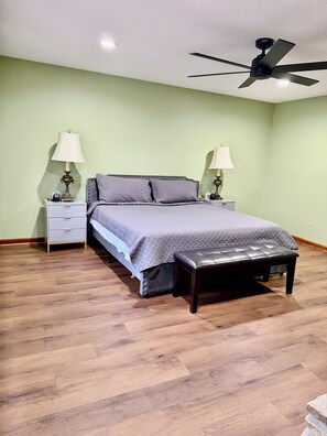 Quarto com cama, duas mesas de cabeceira, um banco, ventilador de teto e piso de madeira.