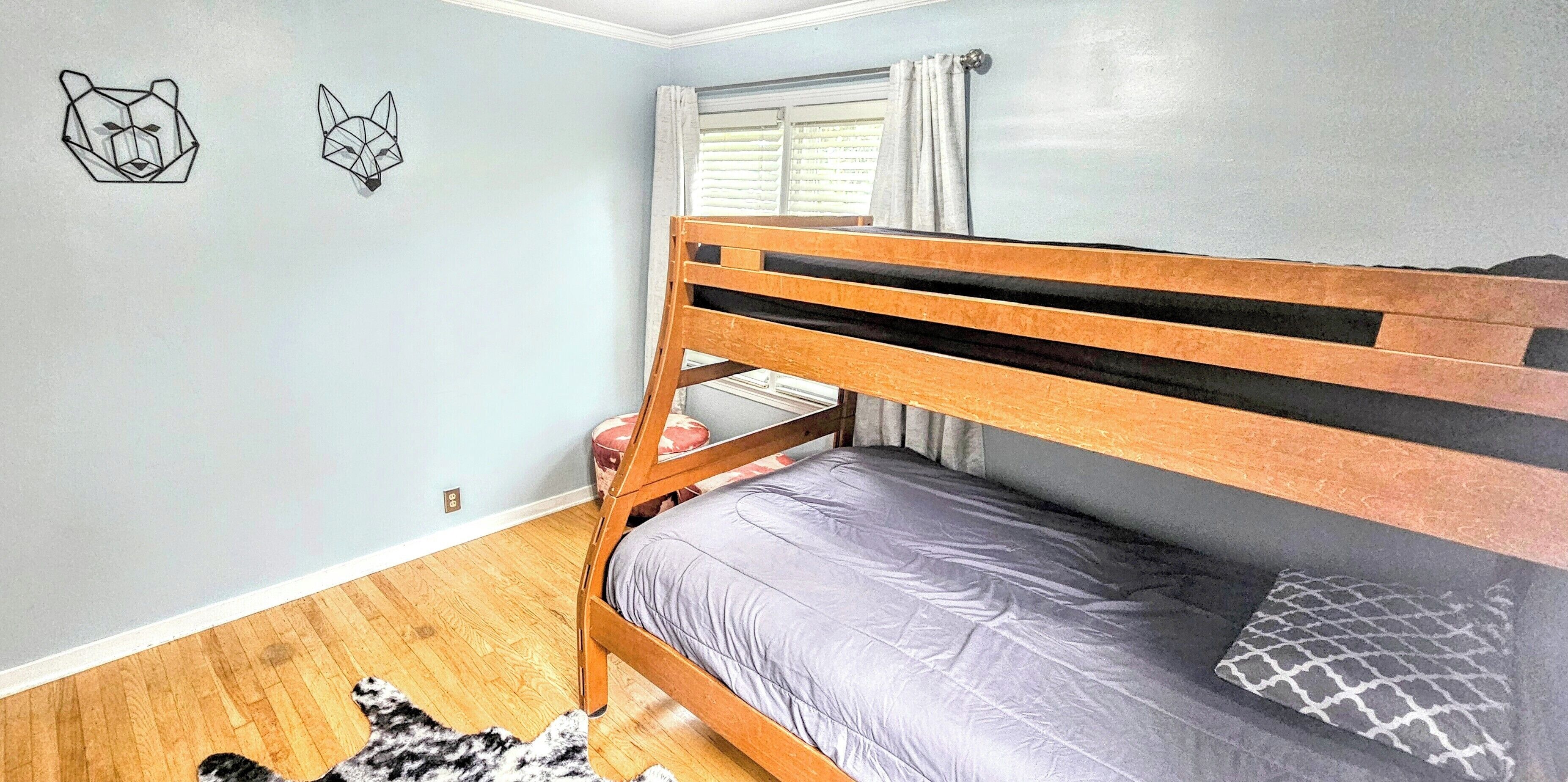 Una cama individual con estructura de madera y colchón gris, un taburete redondo pequeño, un cojín estampado y dos piezas de arte mural geométrico.