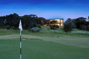 Ein Golfplatz mit Fahnenstange, ein modernes Haus mit großen Fenstern und dichter Baumlandschaft im Hintergrund.